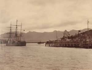 Sepia-toned photograph from 1901 of RRS Discovery departing from its New Zealand base, Lyttelton Harbour, for Antarctica. Masses of well wishers line the wharves to see the ship off.