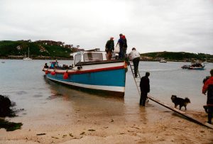 Residents board a small boat using wooden planks and a ladder on the beach. There is no jetty available. 