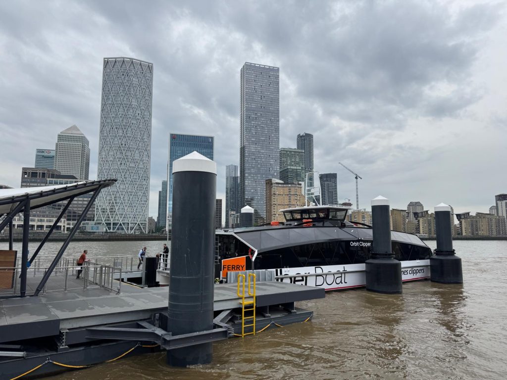 The Orbit Clipper is seen berthed at Rotherhithe pier during berthing trials, with the skyline of Canary Wharf behind. 