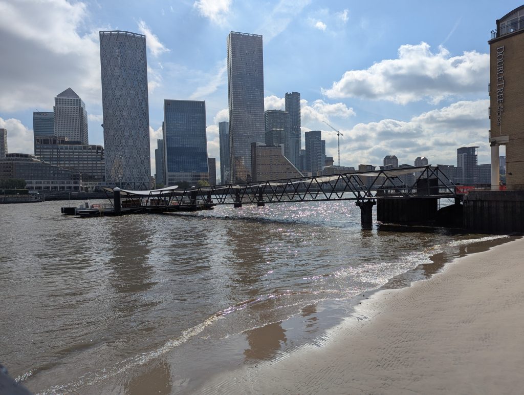 View of the entire new Rotherhithe Pier from the shore on a sunny day. The Canary Wharf skyline is visible behind.  
