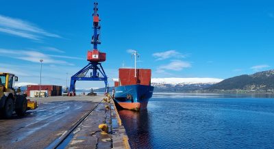 A cargo ship docked at the quayside next to a crane. In the background, there is snow on the hills.